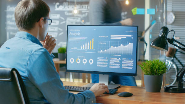 analyst works on a personal computer showing statistics, graphs and charts. in the background his coworker and creative office. - pc imagens e fotografias de stock