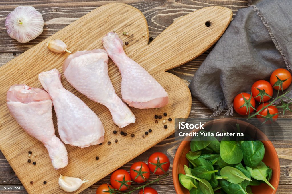 Raw chicken drumstick on a cutting board, cherry tomatoes, spinach, garlic, towel on a wooden table, top view. Raw chicken drumstick on a cutting board, cherry tomatoes, spinach, garlic, towel on a wooden table, top view Bird Stock Photo