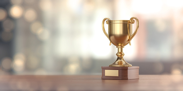 Gold award cup standing on wood surface against pale defocused background.  Panoramic composition with copy space.