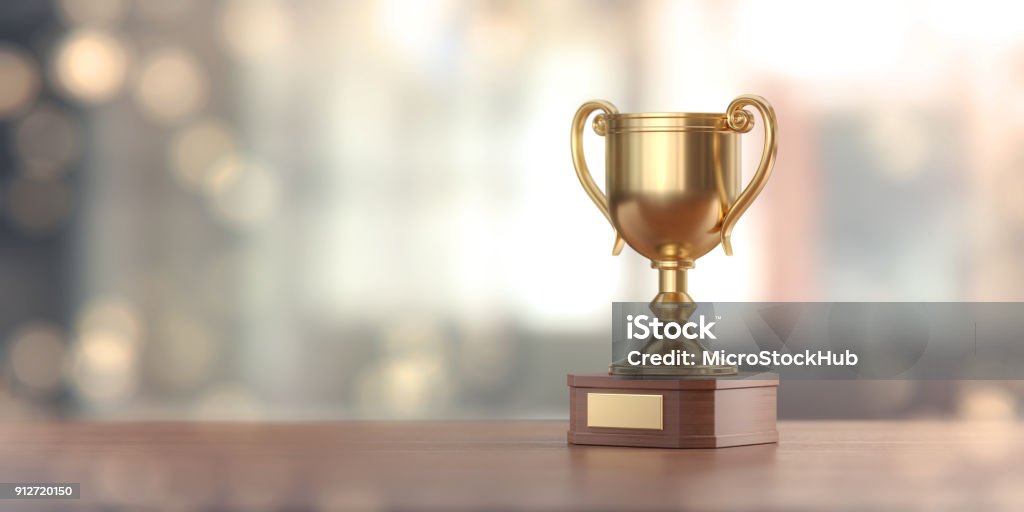 Gold Award Cup Against Defocused Background Gold award cup standing on wood surface against pale defocused background.  Panoramic composition with copy space. Trophy - Award Stock Photo