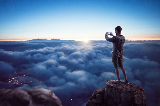 Young man standing on a summit above a scenic cloud cover holding a smartphone in both hands. Night shot with a sun about to rise over the horizon. Through the clouds you can see lights of city underneath.