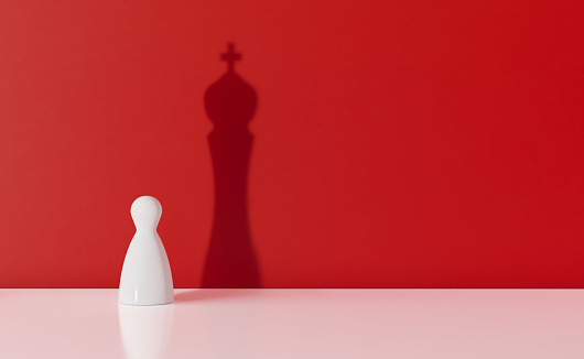 White chess pawn casting the shadow of a king over red background. Leadership and ego concept. Horizontal composition with copy space.