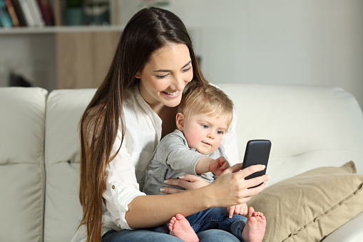 Mother and baby playing with a smart phone sitting on a couch in the living room at home