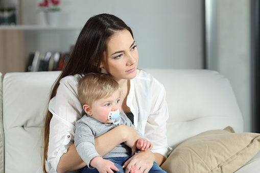 Sad woman looking away with her baby sitting on a couch in the living room at home