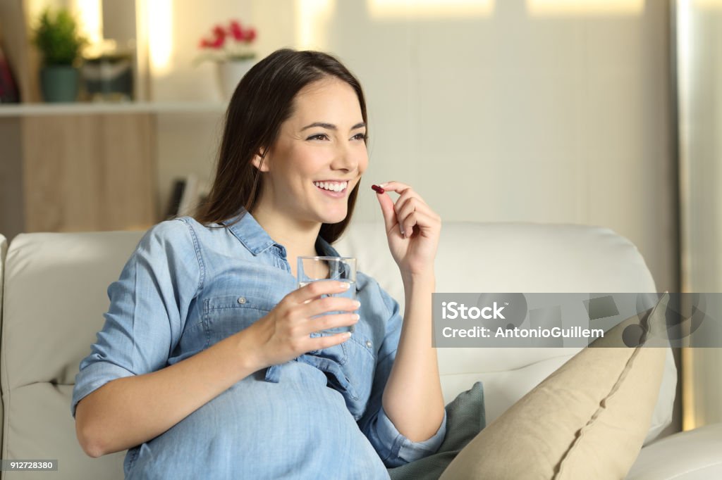 Happy pregnant woman taking a pill at home Portrait of a happy pregnant woman taking a pill sitting on a couch in the living room at home Pregnant Stock Photo