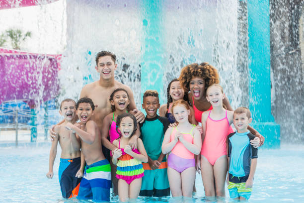 Camp counselors with children at water park A multi-ethnic group of children, 6 to 11 years old, having fun at summer camp at a water park. They are standing with two camp counselors in front of falling water, smiling and laughing, looking at the camera. male swimsuit standing arm around stock pictures, royalty-free photos & images