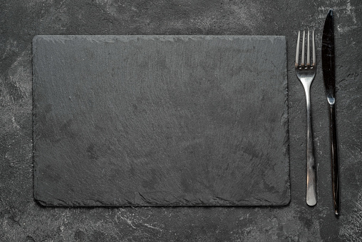 empty black granite stone rectangle board with fork and knife on black textured cement background, top view vith copy space for your text