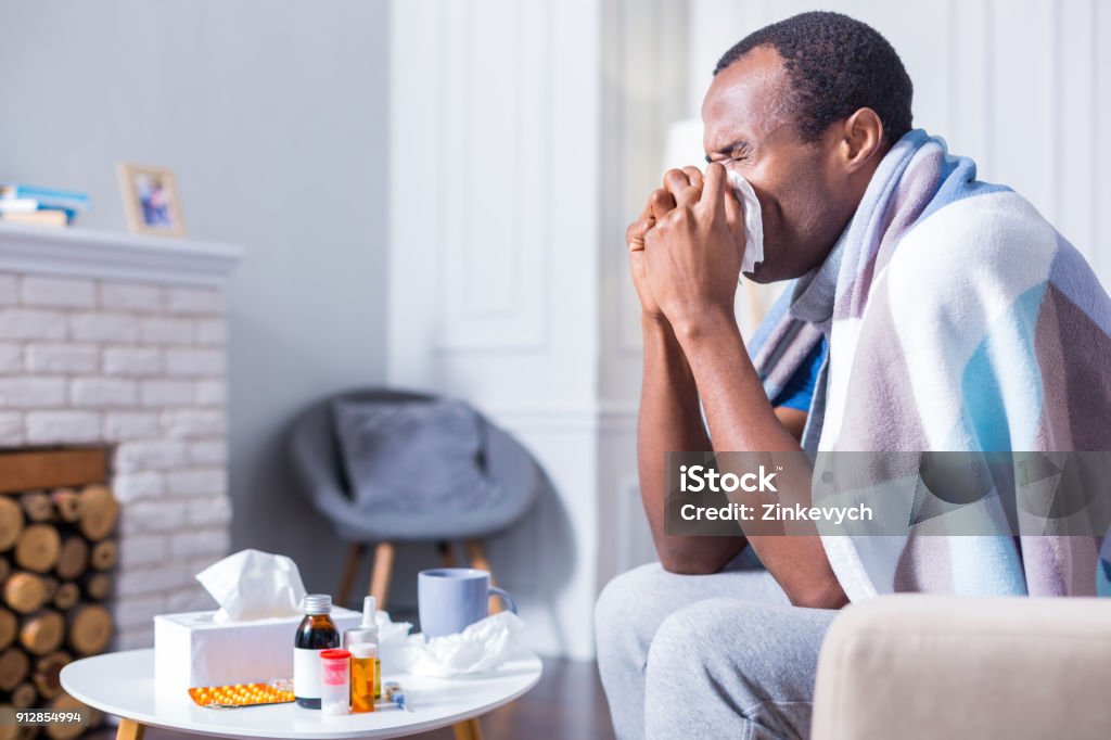 Nice adult man sneezing Seasonal cold. Nice handsome adult man sitting on the sofa and holding a paper tissue while sneezing Cold And Flu Stock Photo