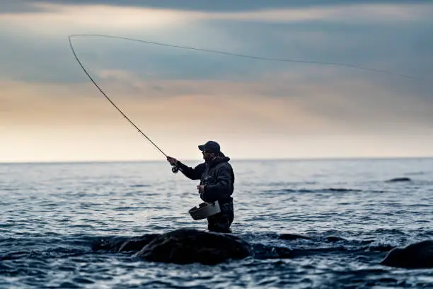 Moody portrait Candid of a fly fisherman waist deep in water casting out his line as he tries to entice a sea trout on to his line. Photographed from the waters edge with a calm sea and a stormy sky. Colour, horizontal with lots of copy space, photographed on the island of Møn in Denmark. The fishermen is wearing green waders and a matching jacket, photographed in a modern contemporary style.