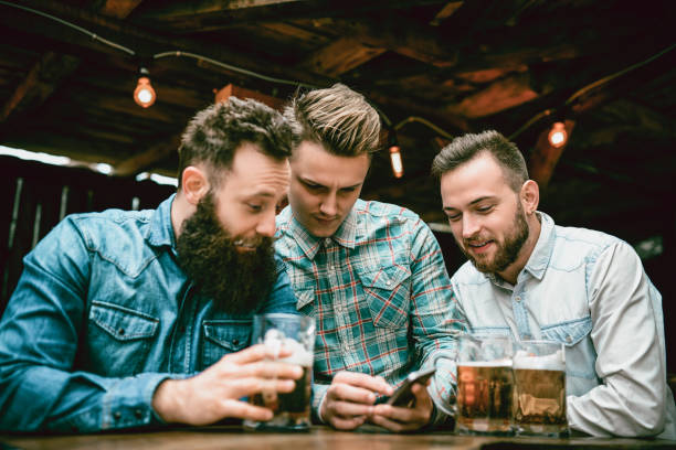 Guys at Pub Drinking Beer and Looking at Smartphone Guys at Pub Drinking Beer Looking at Smartphone and Having a Great Time. friends in bar with phones stock pictures, royalty-free photos & images