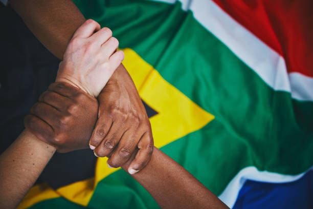 Three multiracial hands are clasped in unity on top of the South African national flag