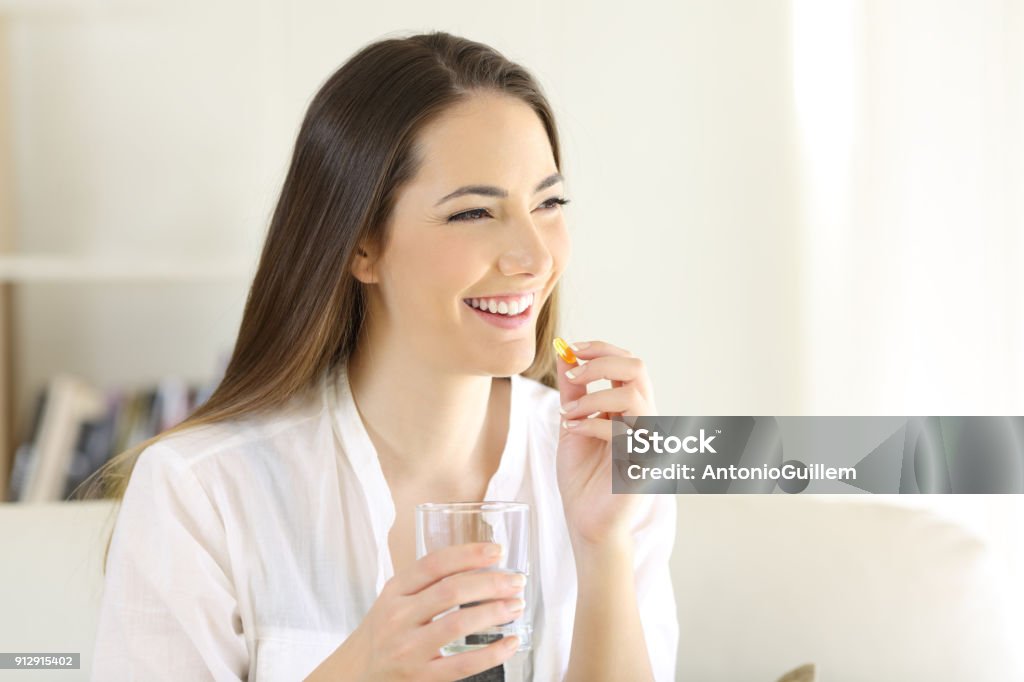 Happy woman taking a vitamin yellow pill at home Happy woman taking a vitamin yellow pill sitting on a couch in the living room at home Capsule - Medicine Stock Photo