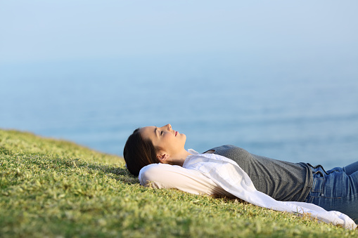 Side view portrait of a relaxed woman resting lying on the grass in the coast with the ocean in the background