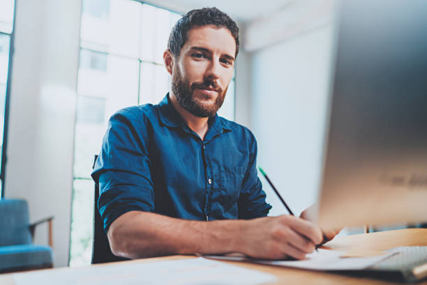 istock Bearded young businessman working at sunny office.Man using contemporary laptop and making notes document.Horizontal.Blurred background. 912952712
