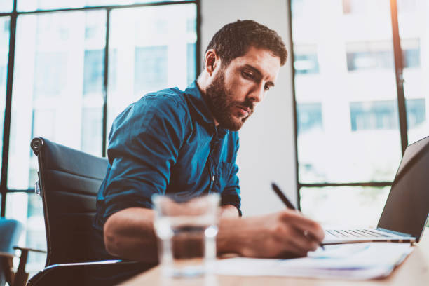istock Young attractive man working at sunny loft office on laptop while sitting at wooden table.Businessman analyze digital reports on notebook computer.Blurred background,horizontal. 912955324