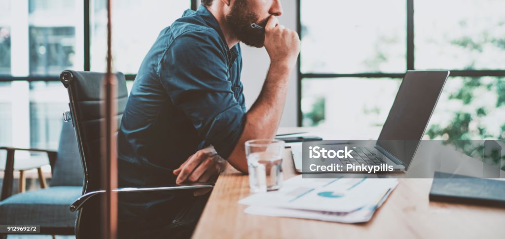 Jeune collègue songeur travaillant au loft ensoleillé lieu de travail tout en étant assis à la table en bois. L’homme analyser le document sur l’écran de l’ordinateur. Arrière-plan flou. Échelle horizontale. - Photo de Bureau - Lieu de travail libre de droits