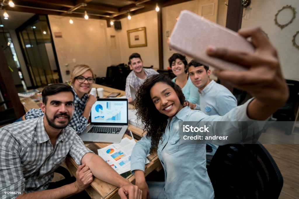 Happy group of coworkers taking a selfie at a creative office Portrait of a happy group of Latin American coworkers taking a selfie at a creative office with a cell phone. **DESIGN ON SCREEN AND DOCUMENTS WERE MADE FROM SCRATCH BY US** Selfie Stock Photo
