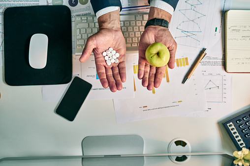 High angle shot of an unrecognizable businessman holding an apple in one hand and a bunch of pills in the other at the office