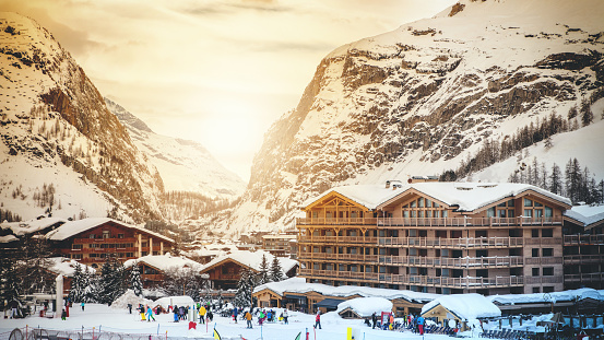 Horizontal color photography of unrecognizable crowd and group of people skiing in famous ski tourist resort of Val d'Isere in European Alps mountains, in Savoie, in Tarentaise mountain range, Auvergne-Rhone-Alpes region in France. People enjoying and skiing winter season in a beautiful landscape with mountains and some wooden chalet, by a sunny january day with bright sunset, using ski lift equipements on ski slopes with deep snow.