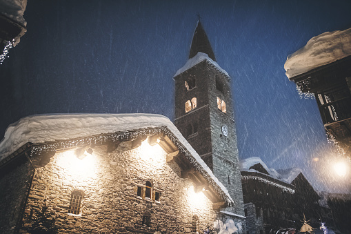 Color horizontal composition photography of bell tower, old steeple stoned church of Val d'isere french ski resort illuminated during a snowy night view from street. This image was taken in winter season in Val d'Isere, a french famous ski resort village in Tarentaise mountains, in Savoie, Auvergne-Rhone-Alpes region in European Alps.