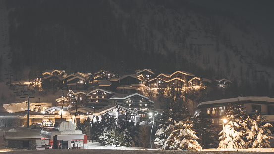Color dark photography of Val d'isere ski resort illuminated village by snowy night in European Alps in winter, with some deep snow and ski lift equipements lost in fog. This image was taken in winter season in Val d'Isere, a french famous ski resort village in Tarentaise mountains, in Savoie, Auvergne-Rhone-Alpes region in European Alps.