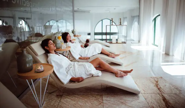 Young hispanic female friends in white robes relaxing together at wellness hotel resort spa