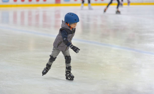 Adorable little boy in winter clothes with protections skating on ice rink stock photo