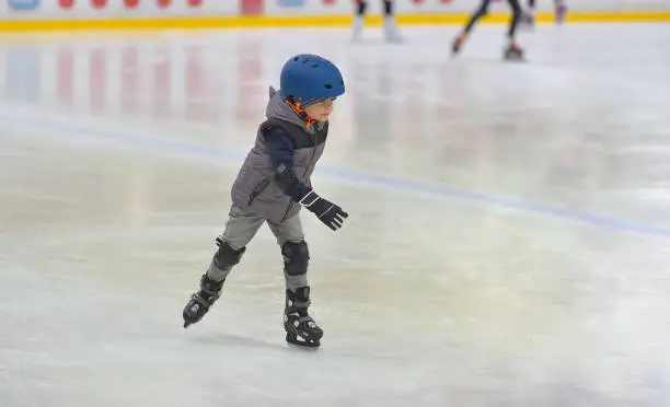 Photo of Adorable little boy in winter clothes with protections skating on ice rink