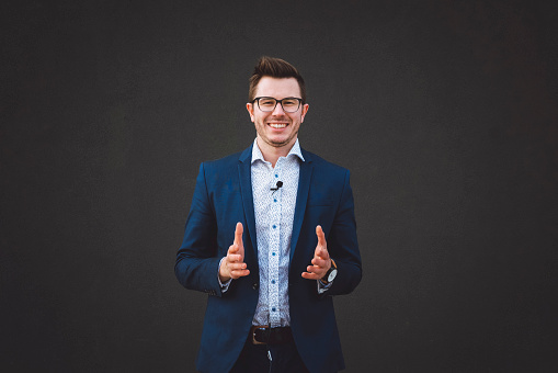 Handsome businessman standing in front of a black wall, posing, smiling, wearing navy blue suit and a shirt with dots on it, wearing glasses, having a nice short hair cut, wearing a watch, businessman approaching his strategies, running business world, young business man feeling happy and confident.