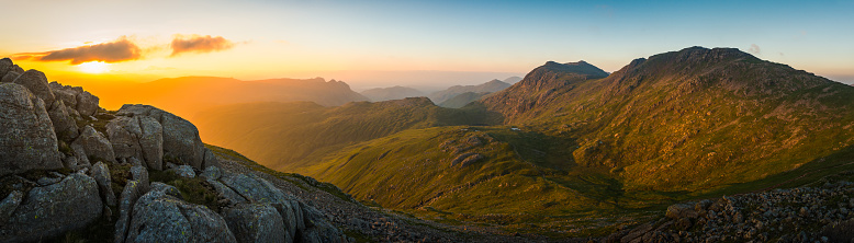 Golden light of sunrise filling the green mountain valleys high in the Lake District National Park overlooked by the iconic rocky summits of the Langdale Pikes and Bow Fell towards Coniston and Windermere, Cumbria, UK.