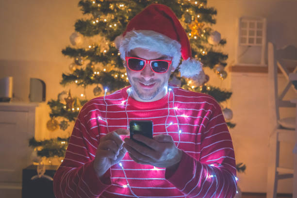 istock Man with santa hat and christmas lights using smartphone. Christmas tree in the background 913179884