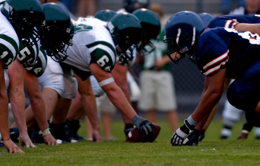 A DSLR of american football players in their football uniforms at the line of scrimmage of a football game. the photo is a close up of the football players with their football helmets, football pads and football gear and a leather football in the green grass at a sporting event. the football field is green grass. and the lighting is natural sunlight during the evening. the image could be titled friday night lights.