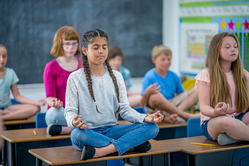 A group of children are indoors in their classroom. They are sitting on their desks and meditating with their legs crossed and eyes closed.