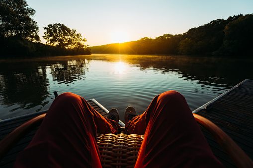 Young Man Sits In Armchair By The Lake And Meets Sunrise Or Sunset, Point Of View. Recration Relaxation Journey Concept