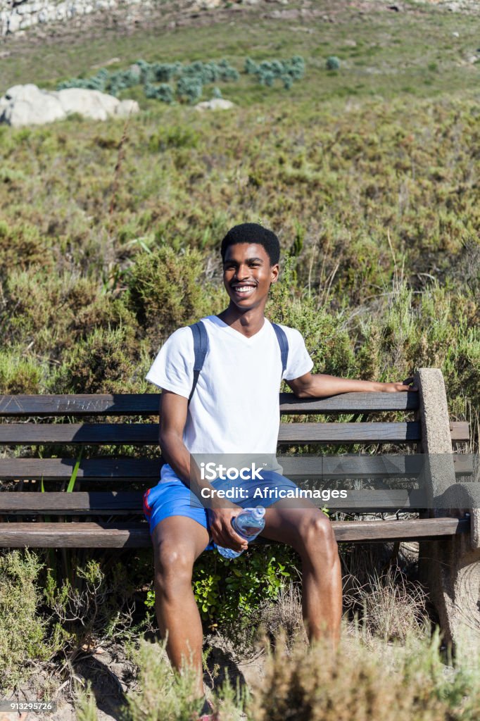 Young man sitting on a bench smiling. Young African male on the mountain sitting on a bench with a big smile on his face. Adult Stock Photo