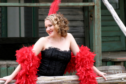 Wild west saloon girl stands outdoors to attract business in a black lace dress with red feather accesories.