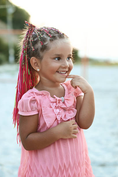 Cute little tanned girl with colored braids hairdo wearing a pink dress is very curious at the summer beach stock photo