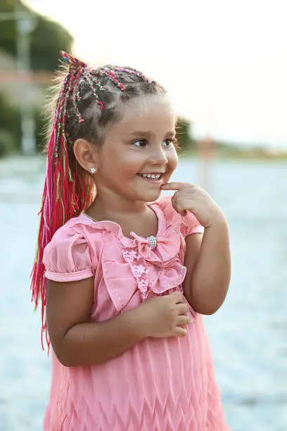 Photo of Cute little tanned girl with colored braids hairdo wearing a pink dress is very curious at the summer beach