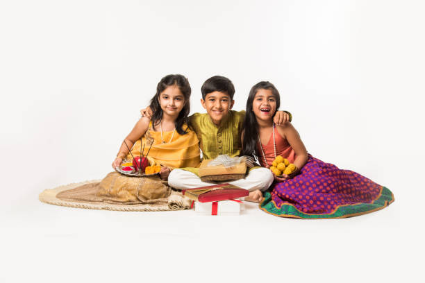 3 indian kids or siblings in traditional wear sitting on sofa or white background, holding gifts and sweets or laddu 3 indian kids or siblings in traditional wear sitting on sofa or white background, holding gifts and sweets or laddu rakhi stock pictures, royalty-free photos & images