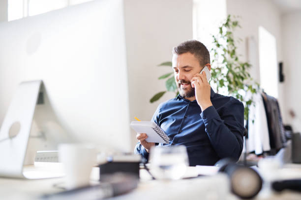 Businessman in wheelchair at the desk in his office. Businessman in wheelchair with smartphone at the desk in the office, making a phone call. ringing stock pictures, royalty-free photos & images