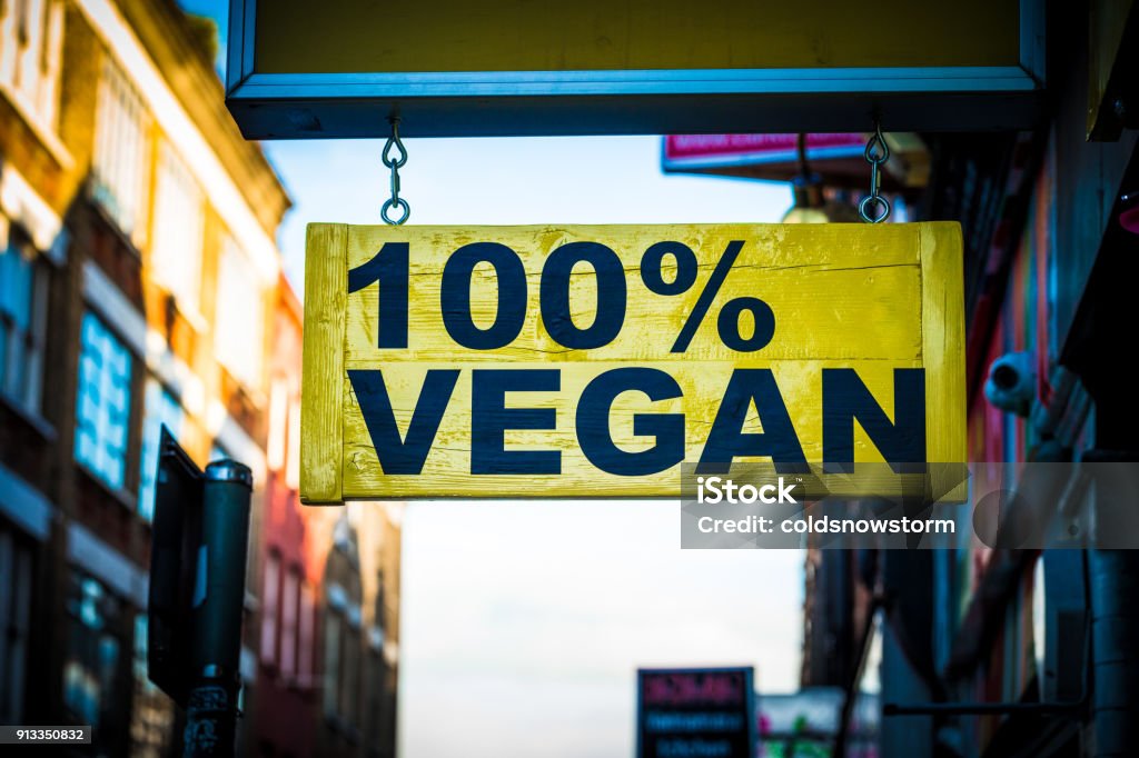 Close up of restaurant sign outdoors on city street saying 100% vegan Close up color image depicting a yellow sign on a city street in London, UK, saying in bold black lettering, 100% vegan. Focus is sharp on the sign in the foreground, while the receding street in the background is blurred out of focus. Lots of room for copy space. Vegan Food Stock Photo