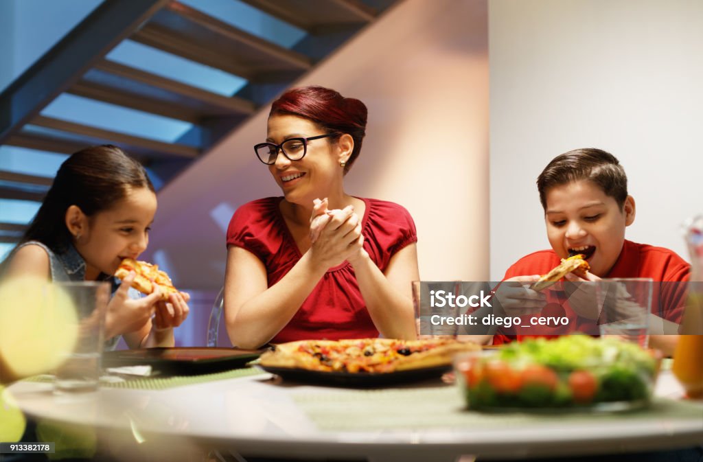 Home Dinner With Happy Family Eating Homemade Pizza Hispanic family with mom, son and daughter having dinner at home and eating homemade pizza. Latino people with mother, boy and girl during meal. Nutrition, food and lifestyle Dinner Stock Photo