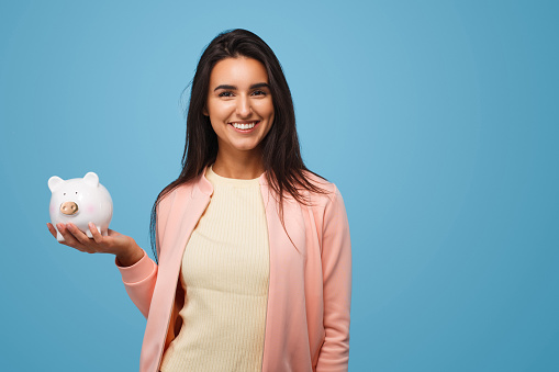 Young smiling woman holding piggy bank on blue backdrop and smiling at camera.