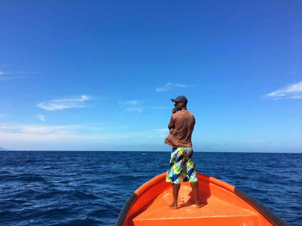 Pensive fisherman on a boat, looking at sea (profile position), sailing in the coastline. Caribbean sea. Afro-caribbean young man. Copy space. Front view. Scenic image. Tropical climate. Sunny day.