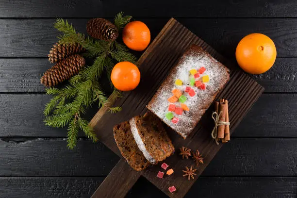 Flatlay of tasty homemade fruit cake with candied fruits, clementines, star anise, cinnamon and fir branch on black background overhead view