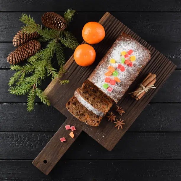Flatlay of chocolate fruit cake decorated with sugar icing, candied fruits, spices and fir branch on dark oak board overhead view