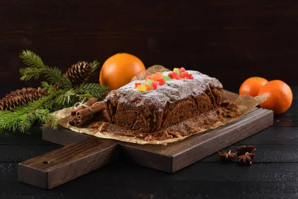 Tasty traditional fruit cake in baking paper decorated with candied fruits, clementines and fir branch on dark background copyspace