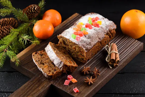 Tasty chocolate fruit cake decorated with candied fruits, clementines, spices and fir branch on carved dark oak board closeup