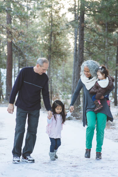 Family Portrait in the Snow Portrait of two beautiful ethnic grandparents going for a walk in the snowy forest with their two grandaughters. Grandma is carrying the younger one and they are holding hands with her big sister. grandparents snow stock pictures, royalty-free photos & images