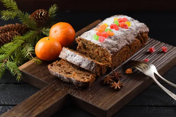 Traditional fruit cake for Christmas served on wooden board with clementines on black background and fur tree
